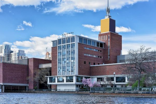 The image shows a red-brick building by a riverside, with a tower and city skyline in the background, under a partly cloudy blue sky.