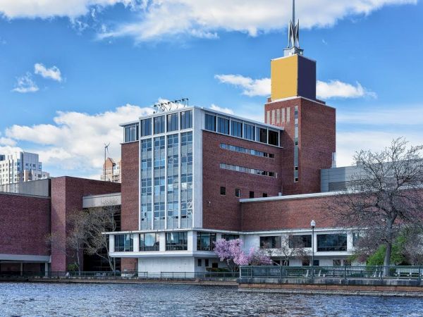 The image shows a red-brick building by a riverside, with a tower and city skyline in the background, under a partly cloudy blue sky.