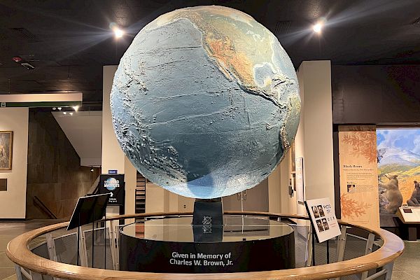 A large globe display in a museum setting, labeled "Given in Memory of Charles W. Brown, Jr." with information panels around it, ending the sentence.