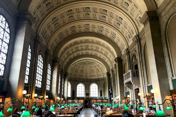 A grand library hall with high arched ceilings, large windows, and wooden desks. Green reading lamps are placed on the desks, and people are studying.