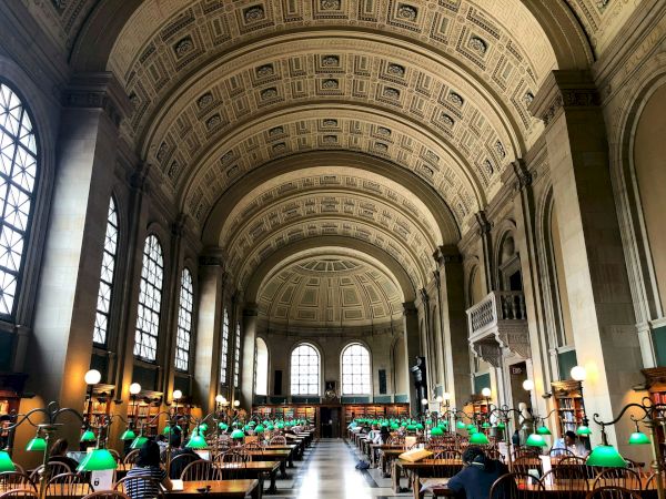 A grand library hall with high arched ceilings, large windows, and wooden desks. Green reading lamps are placed on the desks, and people are studying.