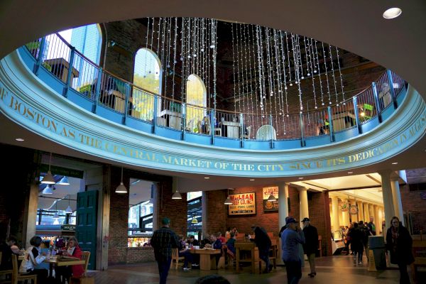 The image shows a bustling indoor market with people dining and walking around, decorative lights hanging from the ceiling and a colorful mezzanine.