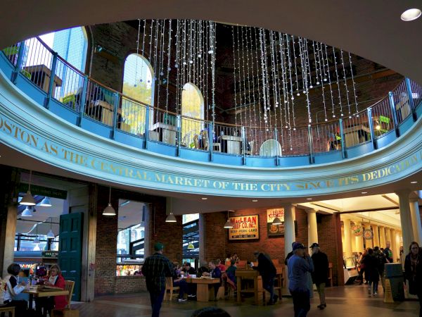 The image shows a bustling indoor market with people dining and walking around, decorative lights hanging from the ceiling and a colorful mezzanine.