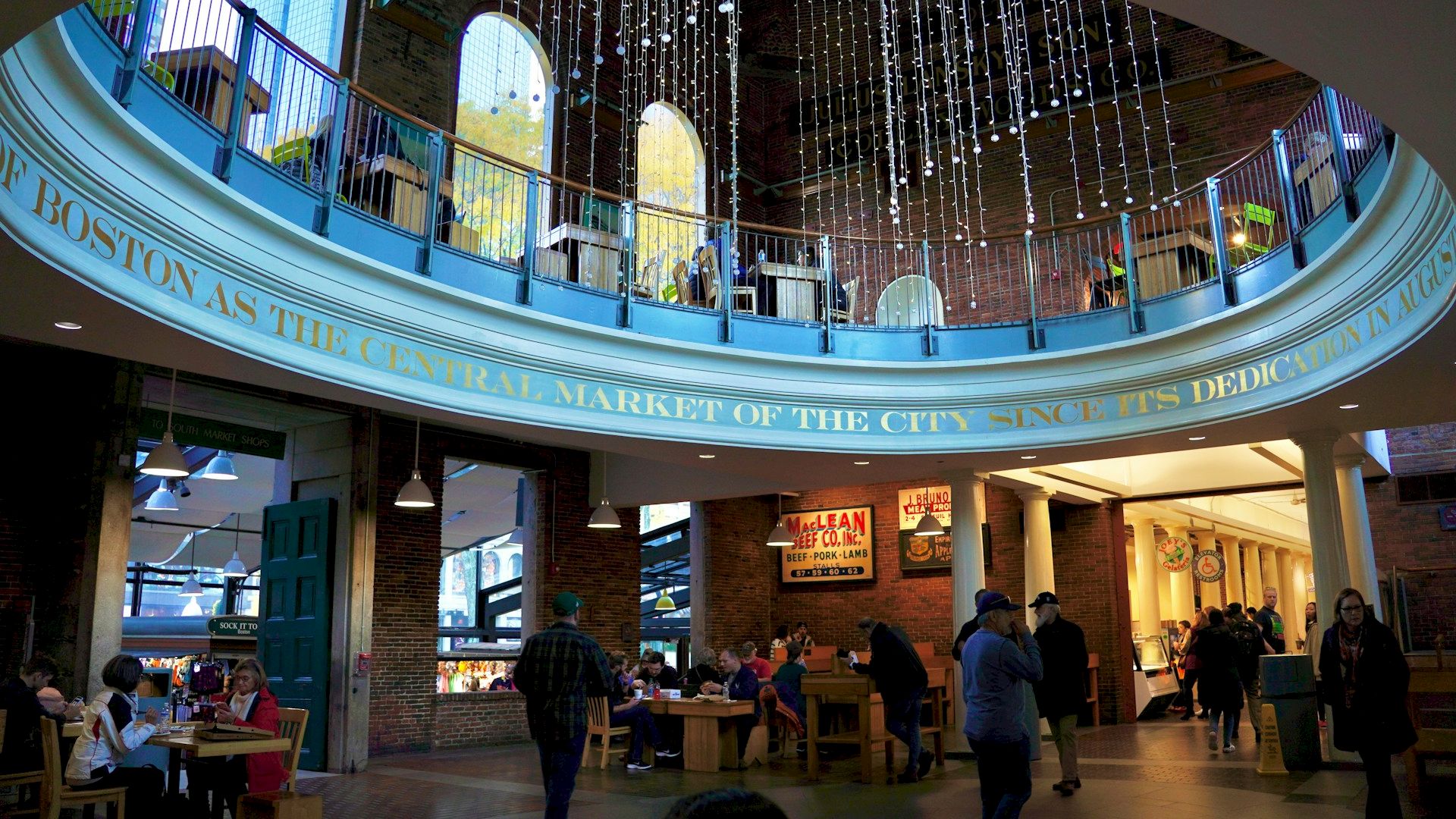Interior view of a bustling public marketplace with people dining at tables, string lights hanging from the ceiling, and various food stalls around.