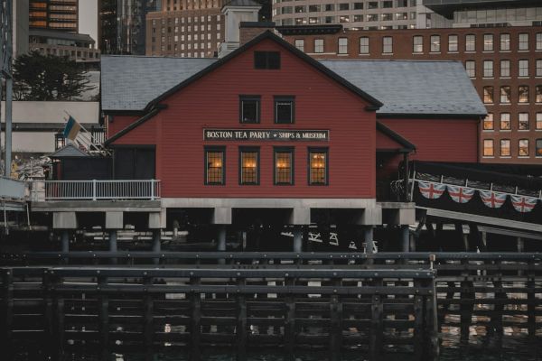 The image shows a red building on a pier, with a reflection in the water, and surrounded by skyscrapers in the background, during dusk.