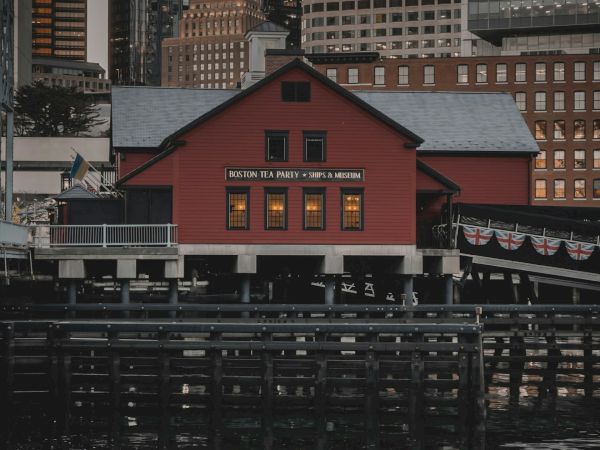 The image shows a red building on a pier, with a reflection in the water, and surrounded by skyscrapers in the background, during dusk.
