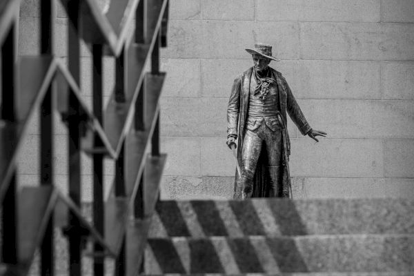 A statue of a man in a hat and long coat stands at the top of a staircase with geometric shadows cast by the railing on the steps.