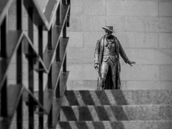 A statue of a man in a hat and long coat stands at the top of a staircase with geometric shadows cast by the railing on the steps.