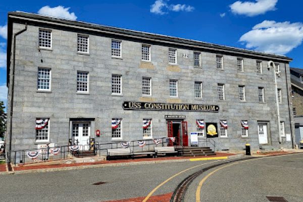 This image shows the USS Constitution Museum, a gray stone building with multiple windows and colorful banners decorating the entrance area.