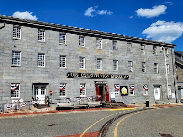 This image shows the USS Constitution Museum, a gray stone building with multiple windows and colorful banners decorating the entrance area.