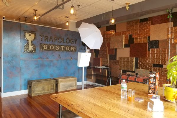 The image shows an indoor setting with rustic decor, branded wall art for "Trapology Boston," wooden furniture, photo equipment, and drinks on a table.