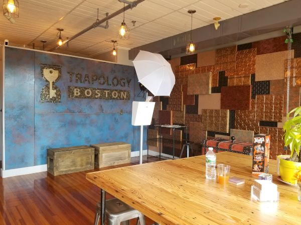 The image shows an indoor setting with rustic decor, branded wall art for "Trapology Boston," wooden furniture, photo equipment, and drinks on a table.