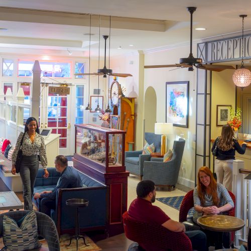 A cozy reception area with people sitting, chatting, and a woman approaching the desk. Stylish decor and warm lighting set the mood.