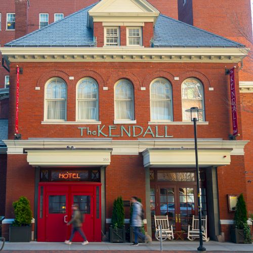 The image shows the exterior of a red brick hotel with the sign "The Kendall Hotel" above the entrance, and people walking by.