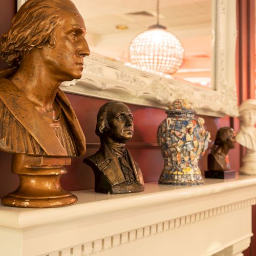 A series of sculptures and a decorated vase are placed on a white mantelpiece with a mirror above, in what appears to be an interior space.