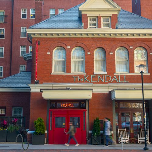 The image shows the facade of "The Kendall" hotel, a red-brick building with arched windows and a sign above the entrance.