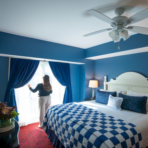 A woman stands by bright curtains in a blue-themed bedroom with a large bed, ceiling fan, and decorative pillows.