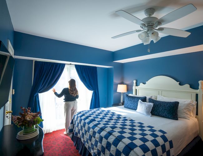 A woman stands by bright curtains in a blue-themed bedroom with a large bed, ceiling fan, and decorative pillows.