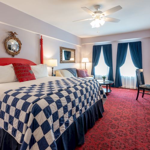 A well-decorated bedroom, featuring a king-sized bed with a checkered quilt, red posts, elegant blue curtains, a ceiling fan, and classic furnishings.