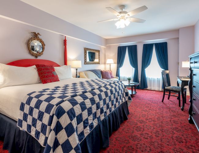 A well-decorated bedroom, featuring a king-sized bed with a checkered quilt, red posts, elegant blue curtains, a ceiling fan, and classic furnishings.