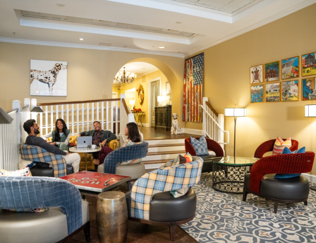 A cozy lounge with people gathered, colorful chairs, wall art, and soft lighting in a warm, inviting setting.