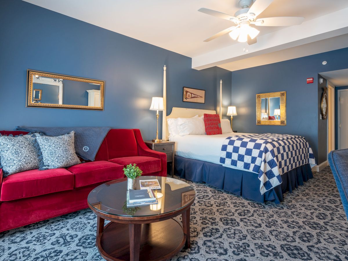 The image shows a hotel room with blue walls, a red couch, a bed with a checkered blanket, a round glass table, and elegant decor throughout the space.