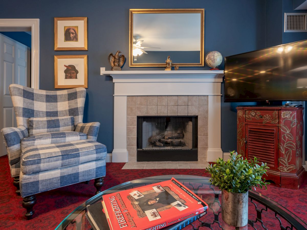 A cozy living room with a plaid armchair, fireplace, TV, and decor including a globe and paintings, with red carpet and a coffee table adorned with books.