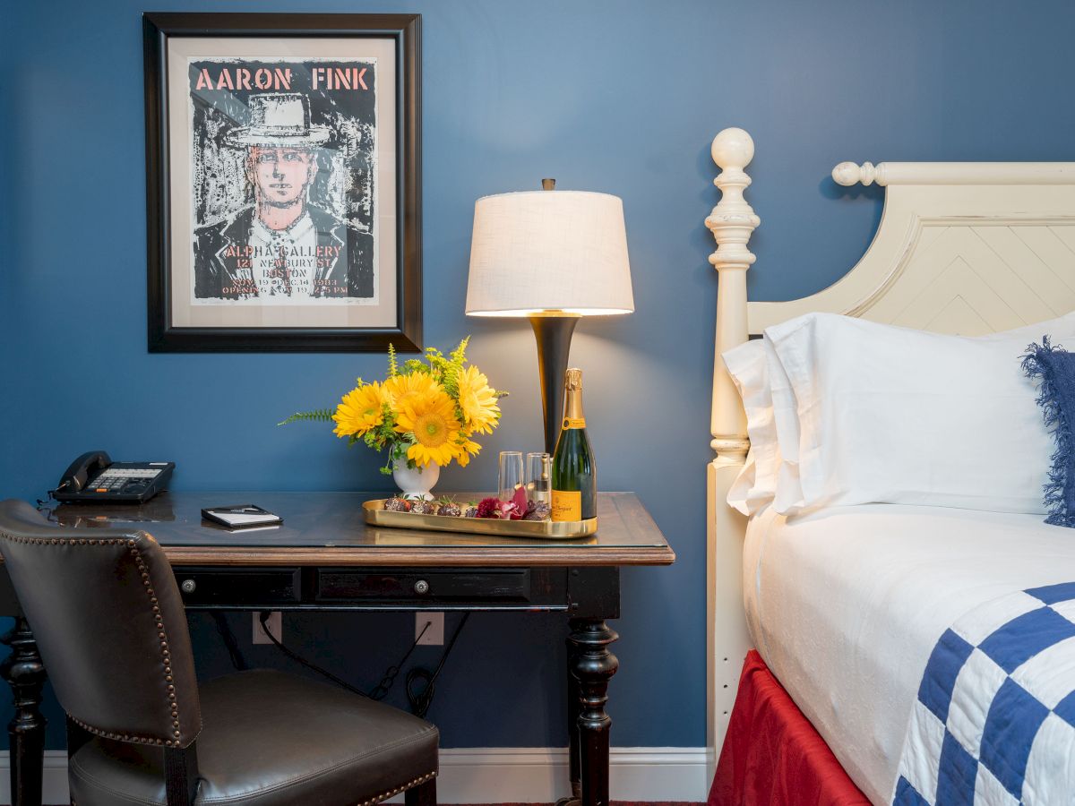 A cozy bedroom with a blue wall, desk with a lamp, flowers, and a champagne bottle beside a bed adorned with a blue and white checkered blanket.