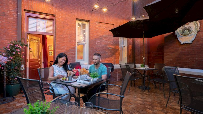 A couple is dining outside at a restaurant patio, with string lights above them and a red brick wall in the background.