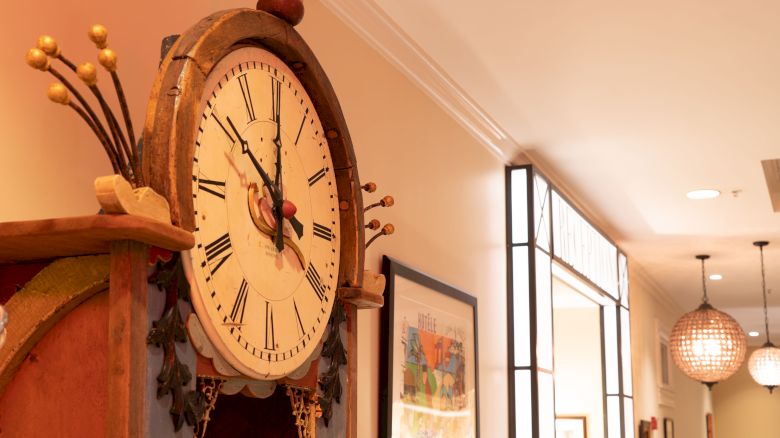 The image shows a decorative clock with Roman numerals, positioned in a well-lit hallway with framed pictures and modern ceiling lights.