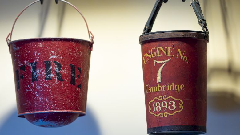 Two vintage red fire buckets, one labeled "FIRE" and the other "ENGINE No. 7 Cambridge 1893," hang from hooks.