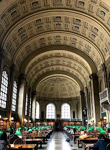 The image shows a grand library hall with a high, ornate ceiling, large arched windows, and rows of tables with green reading lamps.