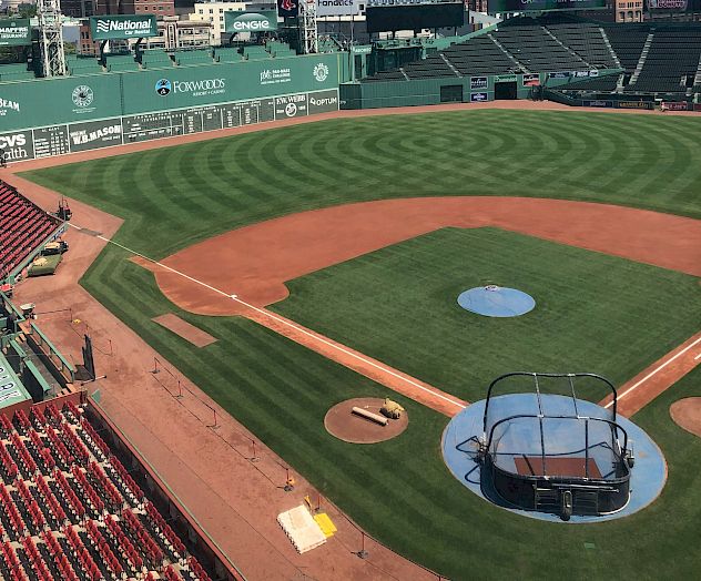 An empty baseball stadium with grass field, dirt infield, stands, and batting cage on home plate. Clear skies with a few clouds are visible.