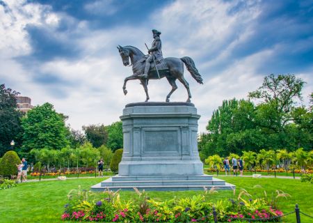 A statue of a rider on a horse stands on a pedestal in a lush garden with colorful flowers under a partly cloudy sky, surrounded by people.