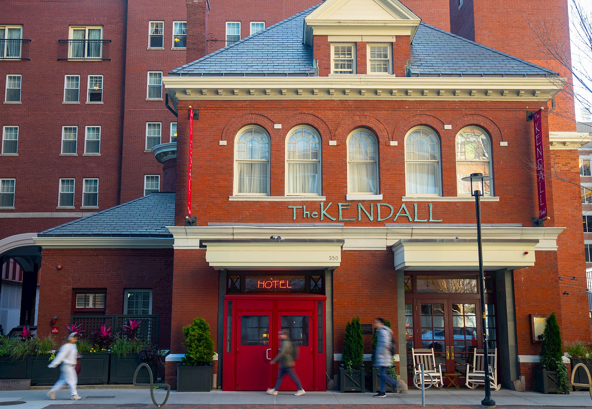 A brick building with "The Kendall" sign, red door, rocking chairs, and people walking past on the sidewalk.