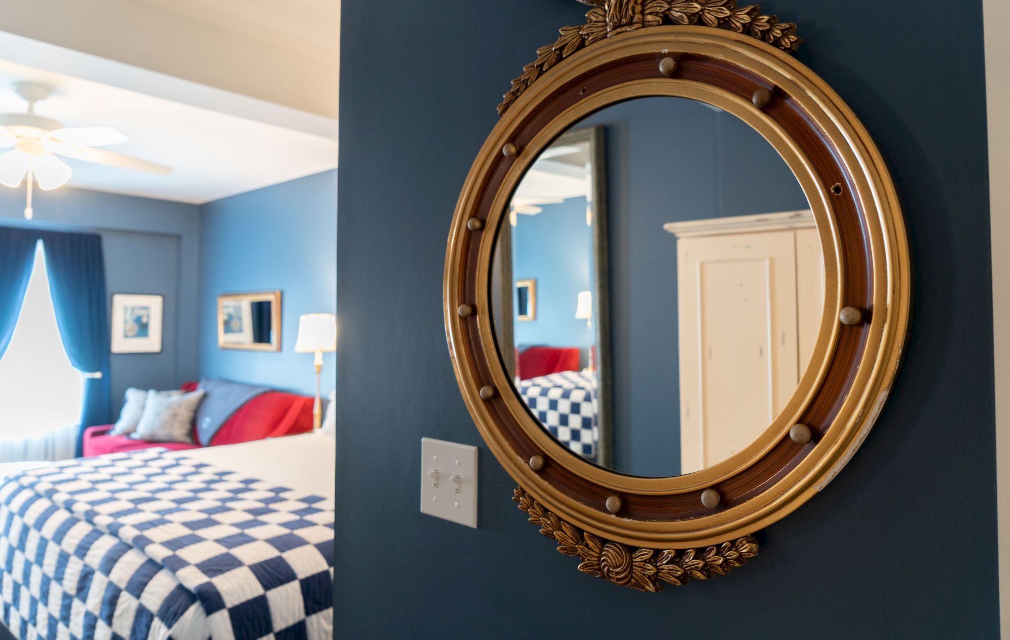 The image shows a bedroom with blue walls, a round decorative mirror, a checkered bedspread, blue curtains, and a white wardrobe.