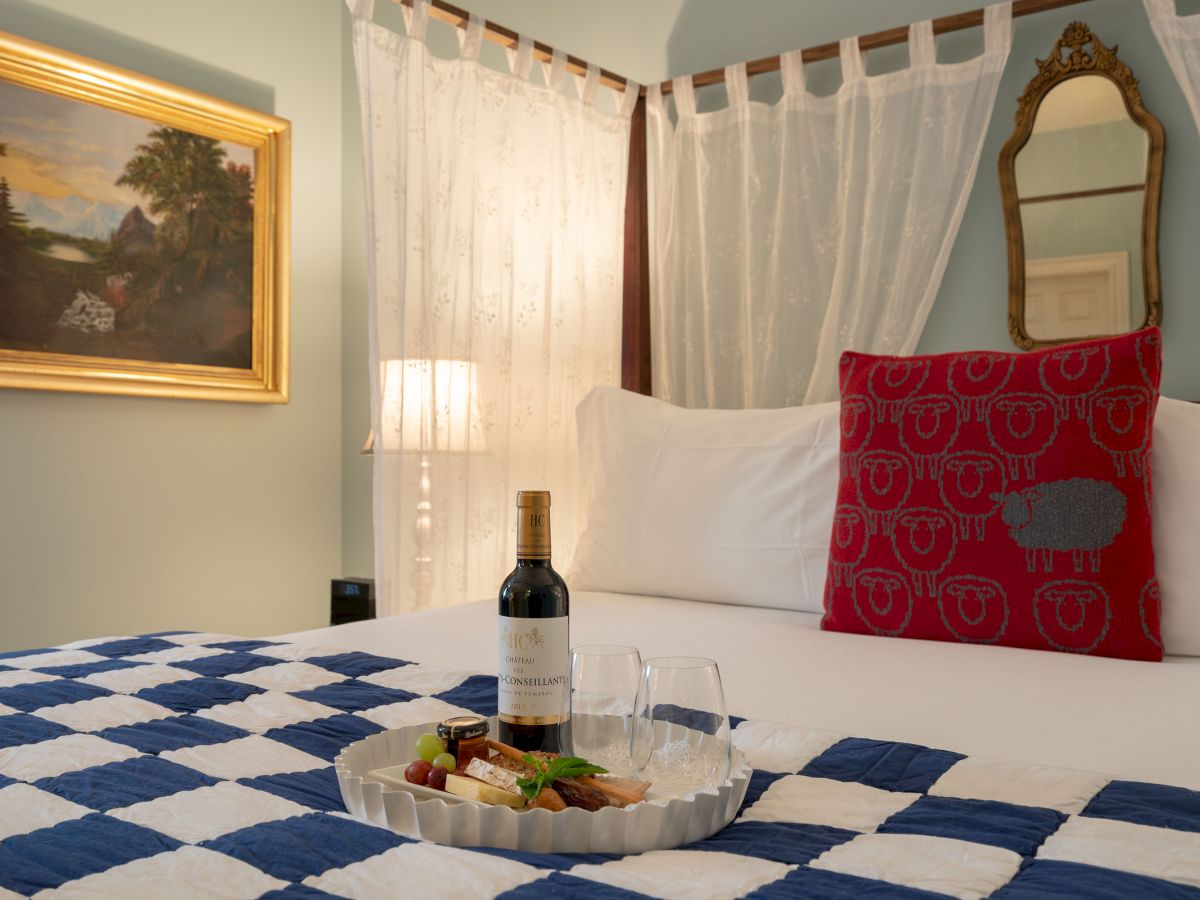 A cozy bedroom features a neatly made bed with a red sheep-patterned pillow and a tray with wine and snacks on a checkered quilt.