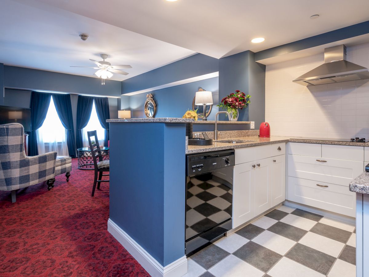 This image shows a modern kitchen leading into a living area with red carpet, blue walls, checkered flooring, and various furnishings.