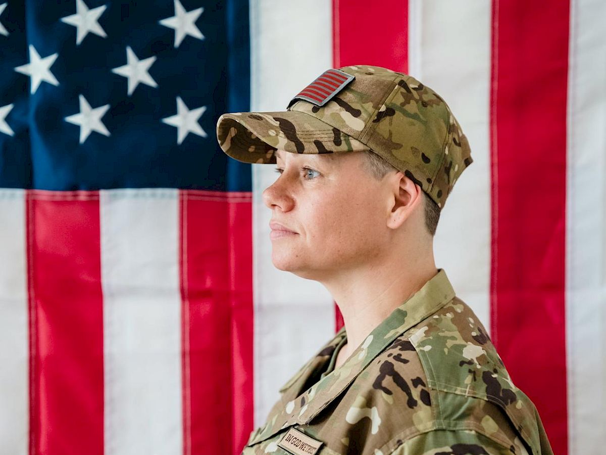 A person dressed in military uniform stands in profile against a backdrop of the American flag.