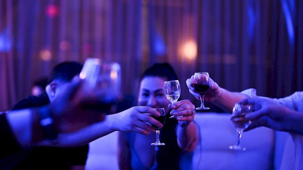 People at a party clink glasses of drinks together under blue and purple lights, celebrating with a toast.