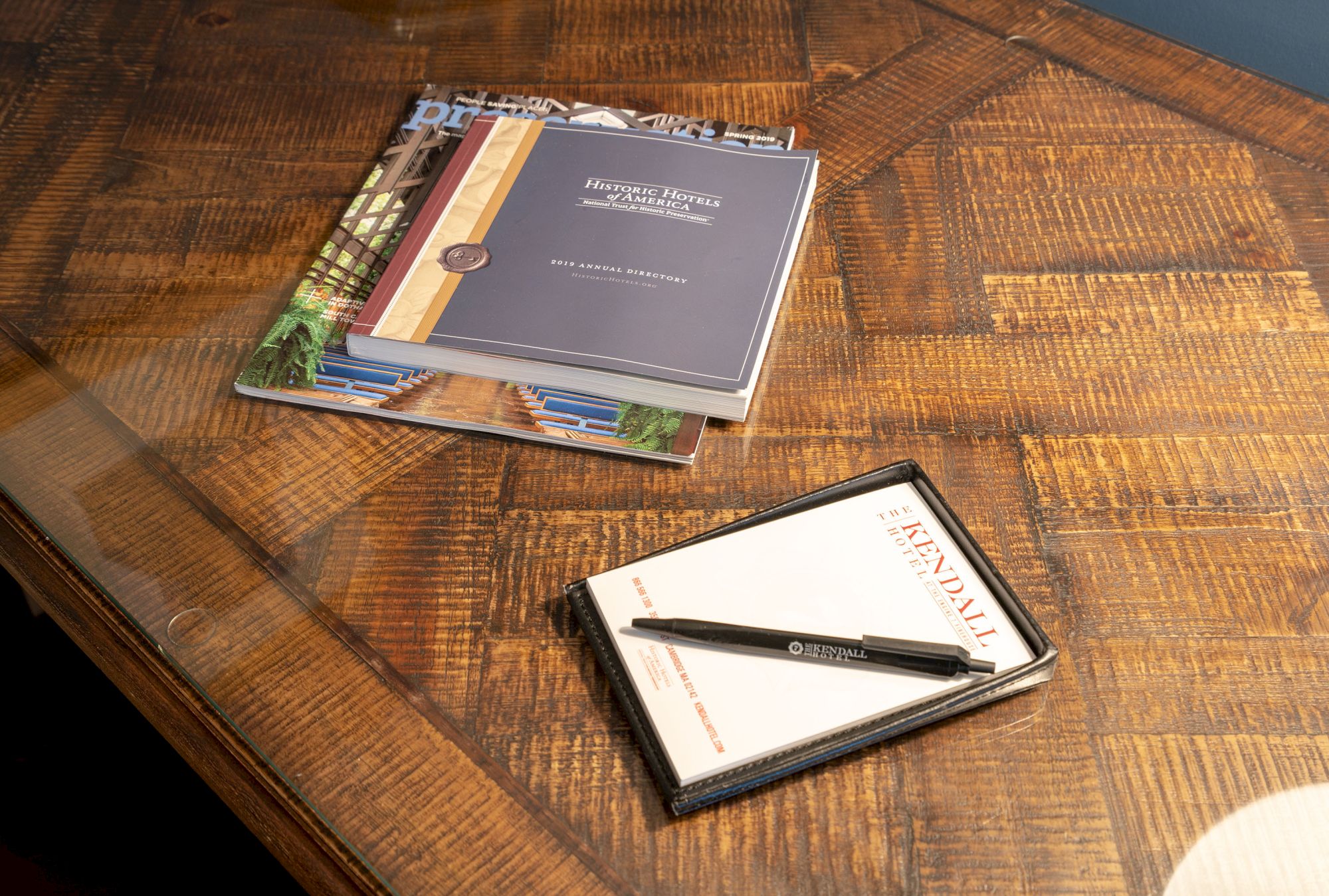 The image shows a wooden table with some brochures and a notepad with a pen placed on top of it, under warm lighting.