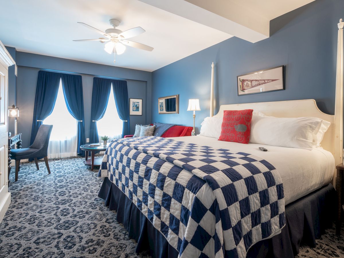 A cozy bedroom with a blue theme, featuring a large bed with a checkered quilt, seating area, and elegant curtains, illuminated by soft lighting.