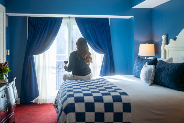 A person sits on a bed in a blue-themed room, holding a glass of wine, gazing out a window with blue curtains.