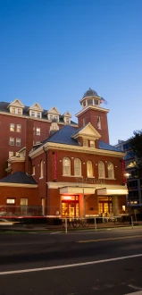The image shows a historic-style brick building with arched windows and a tower, taken during twilight with light trails on the street in front.