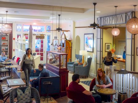 A cozy hotel lobby with people sitting, a reception desk, and shelves with decorations. A mix of seating and warm lighting enhances the space.