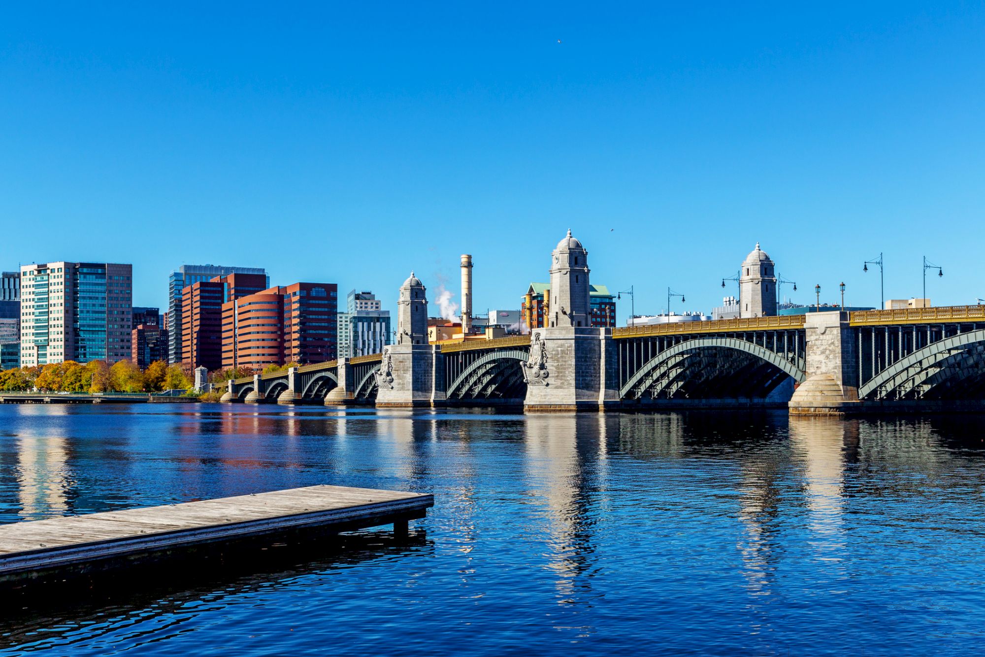 A view of a bridge spanning a river with city buildings in the background and a clear blue sky above, reflecting on the water below.