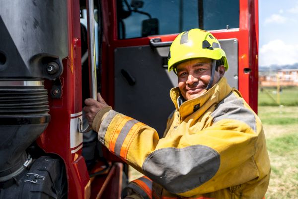 A firefighter in uniform and a yellow helmet smiles while holding onto the door of a fire truck, set in an outdoor location.