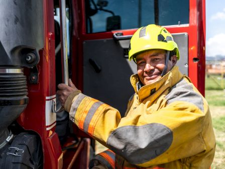 A firefighter in uniform and a yellow helmet smiles while holding onto the door of a fire truck, set in an outdoor location.