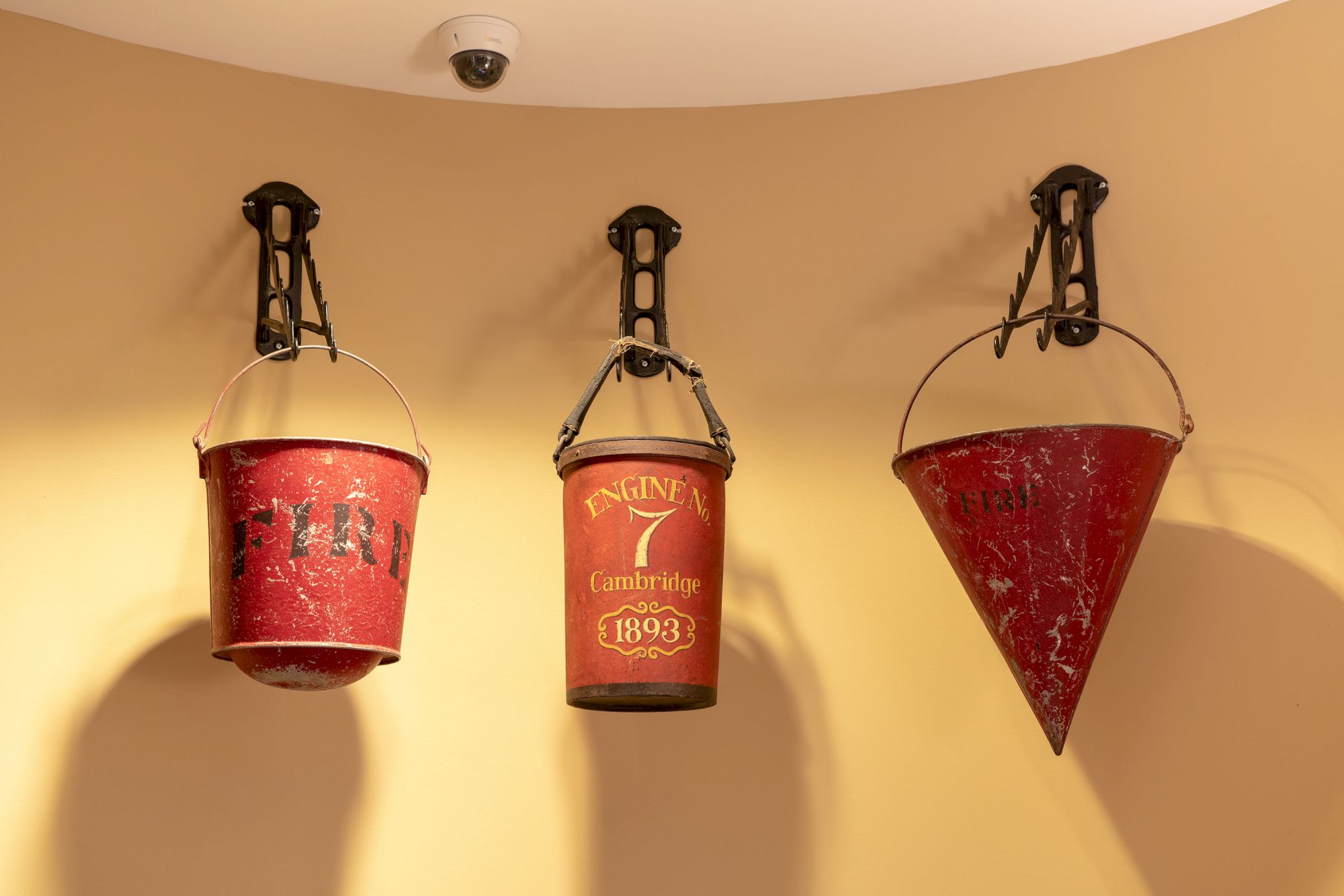 Three vintage fire buckets, including one conical, are mounted on decorative wall brackets under a security camera.