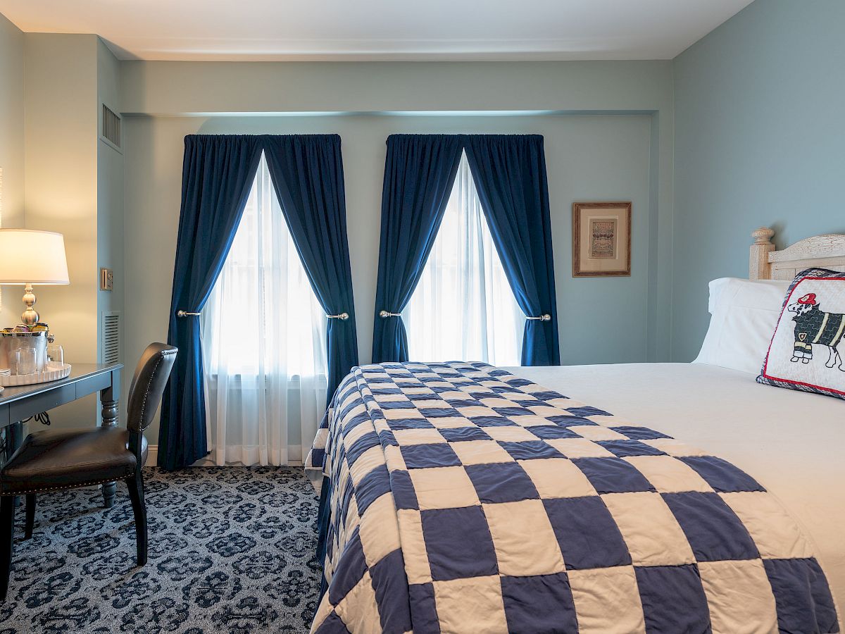 A cozy bedroom with a large bed covered with a blue and white quilt, a desk with a lamp and chair, and two windows draped in navy blue curtains.